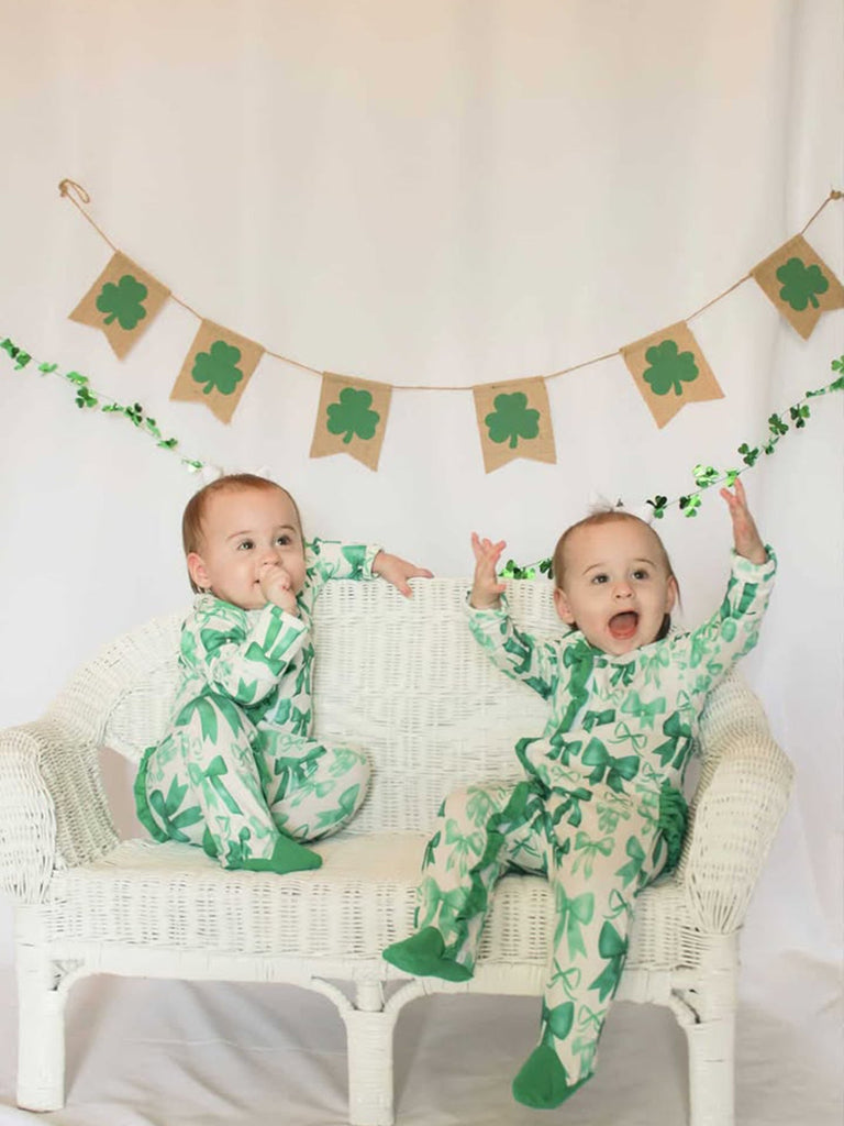 Two babies in green and white outfits with shamrock patterns sitting on a white couch, surrounded by St. Patrick's Day decorations.