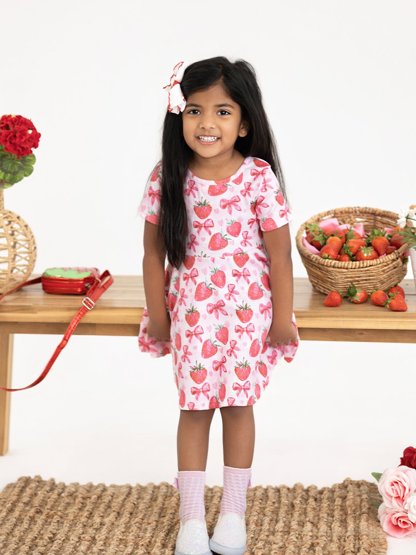 Young girl wearing a strawberry-patterned dress standing in front of a table with strawberries and flowers.