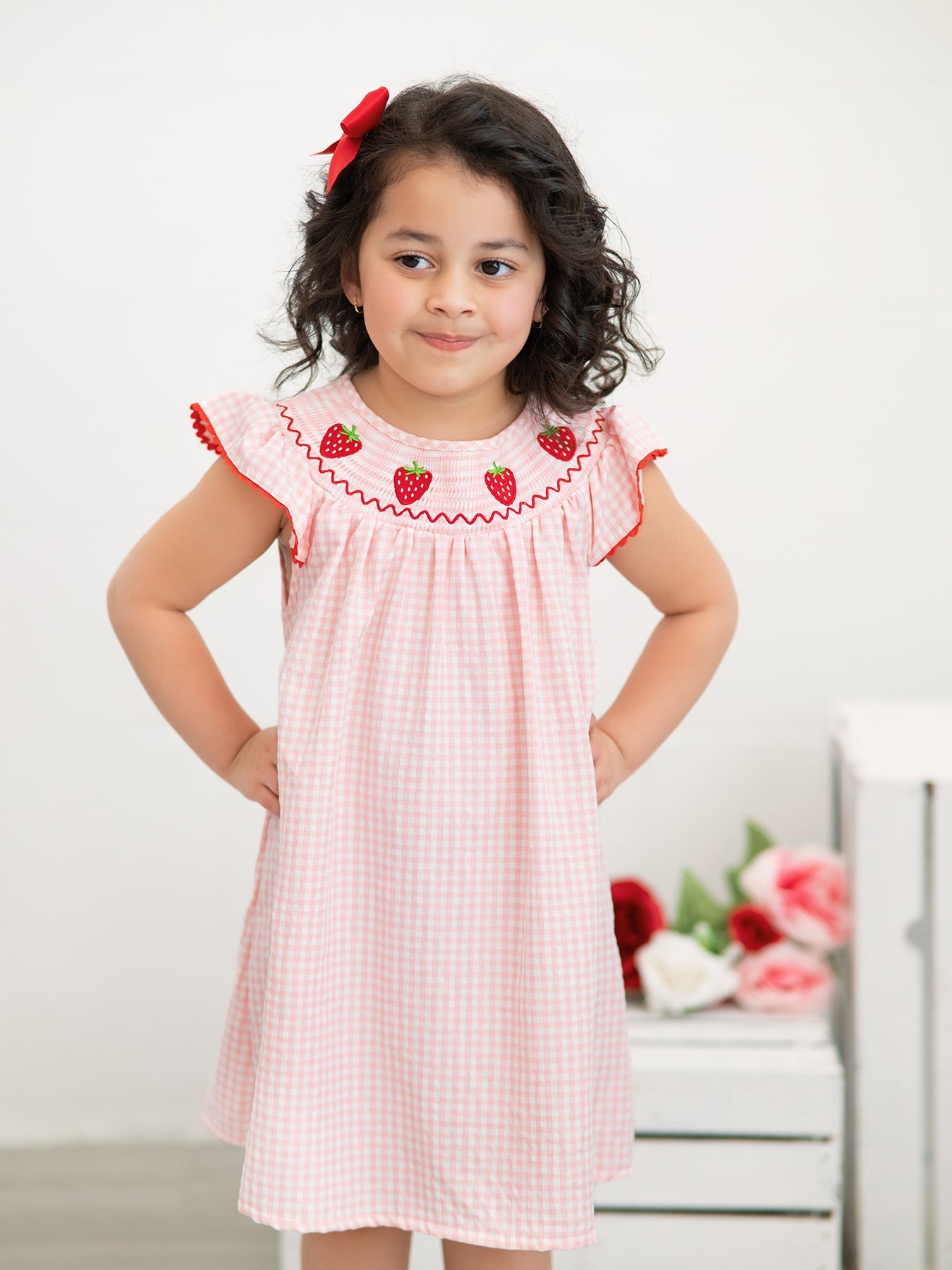 Young girl wearing a pink checkered dress with red embroidery, standing against a white background.