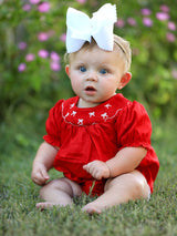 Baby in a red outfit with a large white bow sitting on grass.