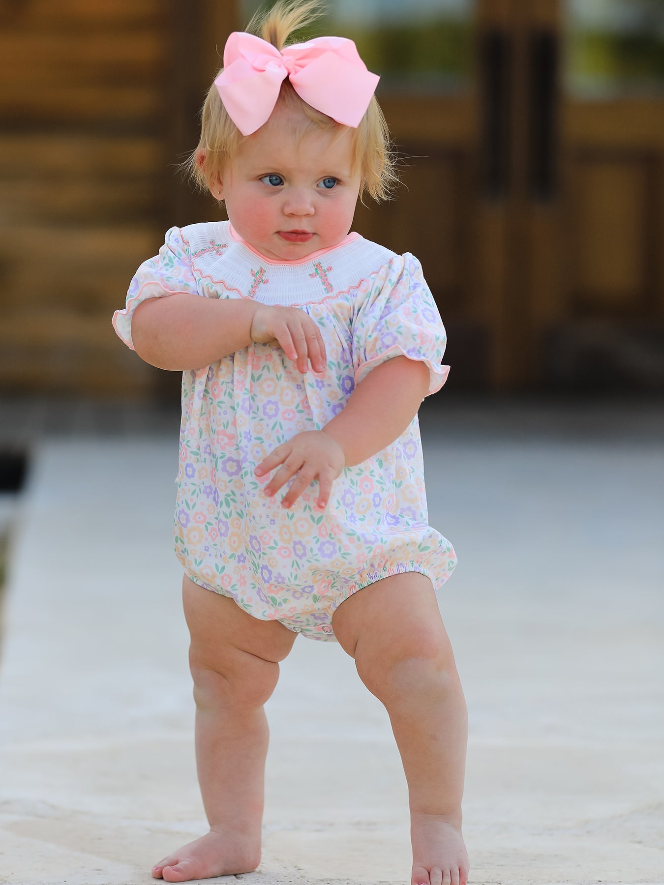 Baby in a floral outfit with a pink bow standing on a sandy surface.