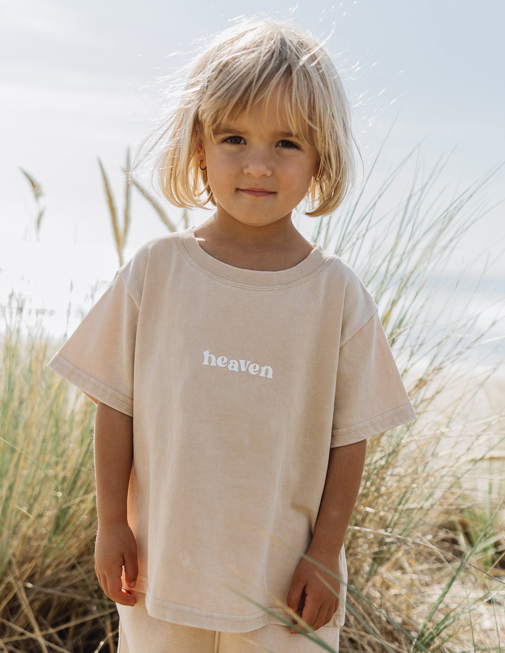 Child wearing a beige 'heaven' t-shirt standing in tall grass.