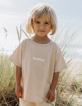 Child wearing a beige 'heaven' t-shirt standing in tall grass.