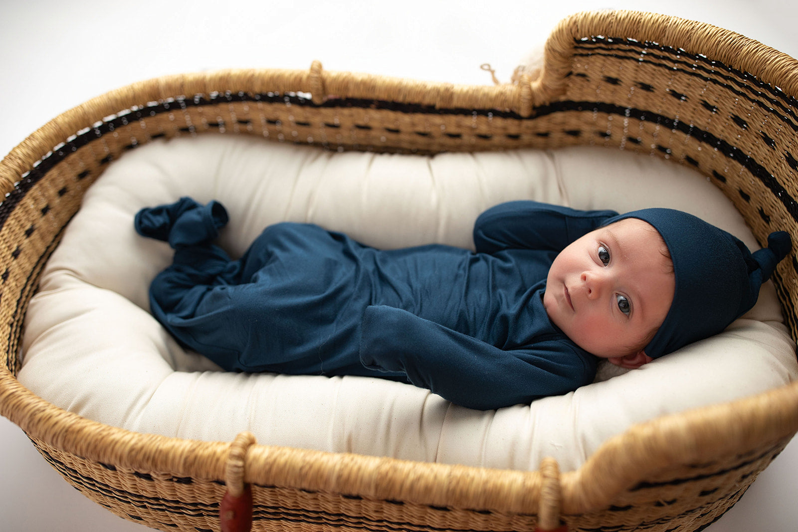 Three Little Tots Navy Blue Knotted Baby Gown. Ultra soft number 1 knotted newborn baby gown. Newborn photoshoot idea or for a pregnancy announcement. Newborn baby in a Snuggle Me Organics in a Plum + Sparrow Moses Basket v
