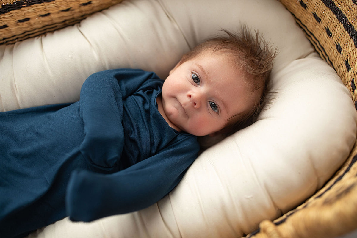 Three Little Tots Navy Blue Knotted Baby Gown. Ultra soft number 1 knotted newborn baby gown. Newborn photoshoot idea or for a pregnancy announcement. Newborn baby in a Snuggle Me Organics in a Plum + Sparrow Moses Basket v