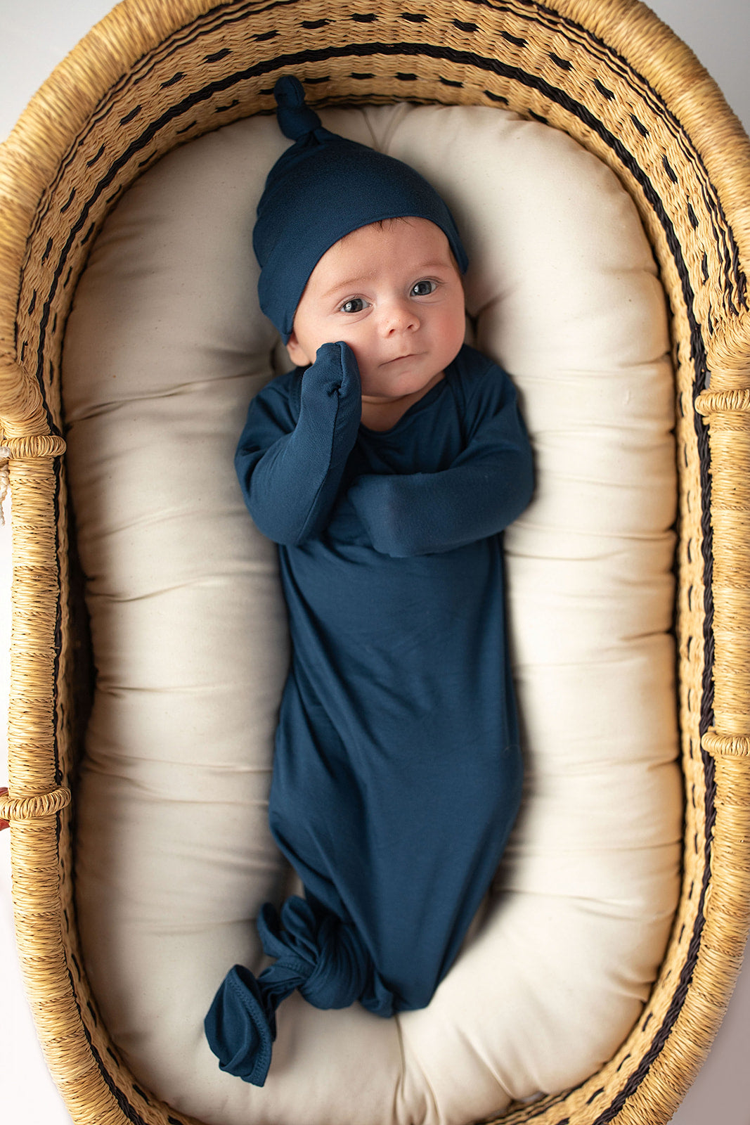 Three Little Tots Navy Blue Knotted Baby Gown. Ultra soft number 1 knotted newborn baby gown. Newborn photoshoot idea or for a pregnancy announcement. Newborn baby in a Snuggle Me Organics in a Plum + Sparrow Moses Basket 