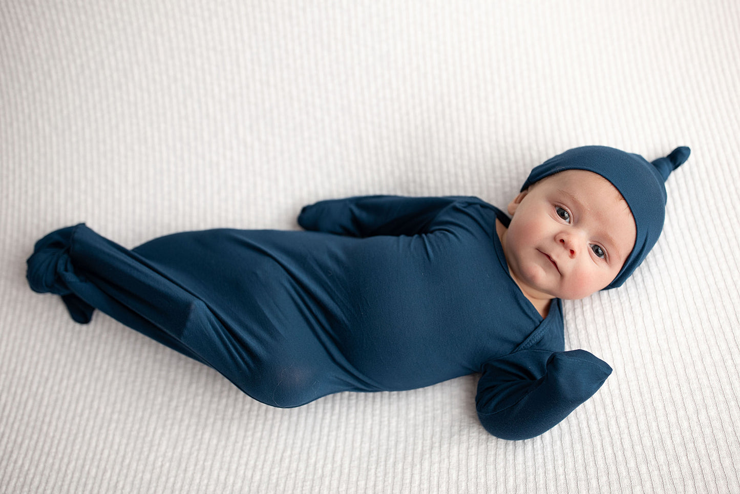 Three Little Tots Navy Blue Knotted Baby Gown. Ultra soft number 1 knotted newborn baby gown. Newborn photoshoot idea or for a pregnancy announcement. Newborn baby in a Snuggle Me Organics in a Plum + Sparrow Moses Basket 