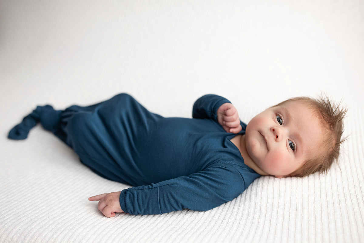 Three Little Tots Navy Blue Knotted Baby Gown. Ultra soft number 1 knotted newborn baby gown. Newborn photoshoot idea or for a pregnancy announcement. Newborn baby in a Snuggle Me Organics in a Plum + Sparrow Moses Basket 