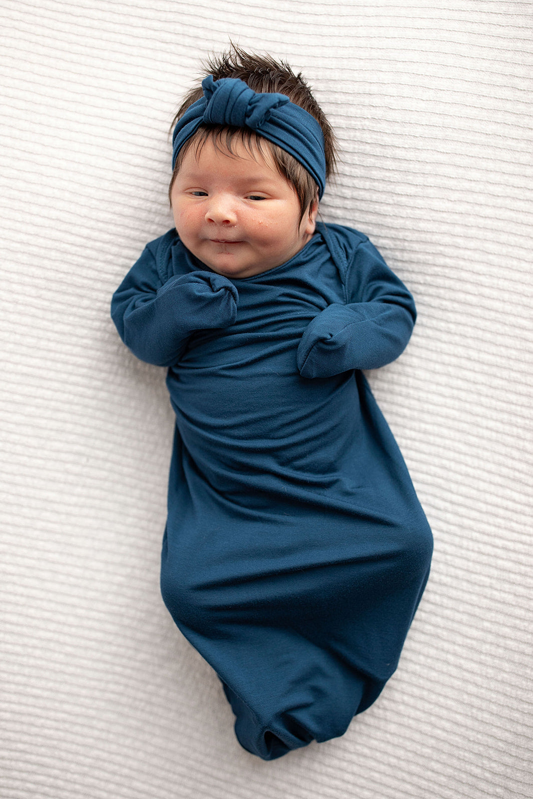 Three Little Tots Navy Blue Knotted Baby Gown. Ultra soft number 1 knotted newborn baby gown. Newborn photoshoot idea or for a pregnancy announcement. Newborn baby in a Snuggle Me Organics in a Plum + Sparrow Moses Basket 