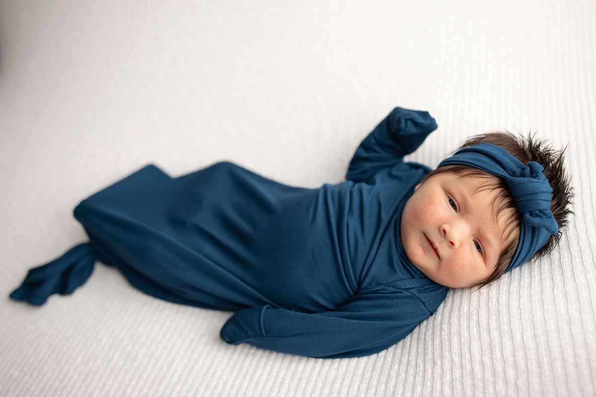 Three Little Tots Navy Blue Knotted Baby Gown. Ultra soft number 1 knotted newborn baby gown. Newborn photoshoot idea or for a pregnancy announcement. Newborn baby in a Snuggle Me Organics in a Plum + Sparrow Moses Basket 