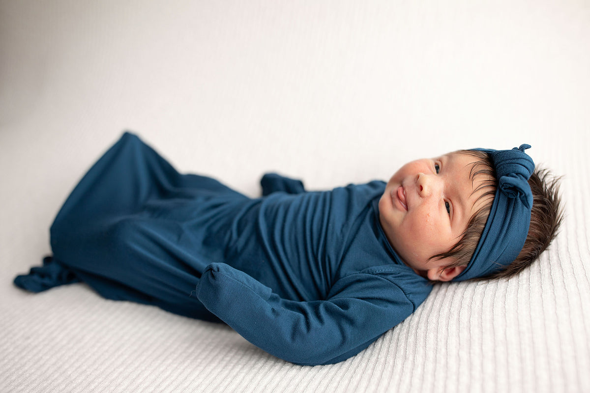 Three Little Tots Navy Blue Knotted Baby Gown. Ultra soft number 1 knotted newborn baby gown. Newborn photoshoot idea or for a pregnancy announcement. Newborn baby in a Snuggle Me Organics in a Plum + Sparrow Moses Basket 