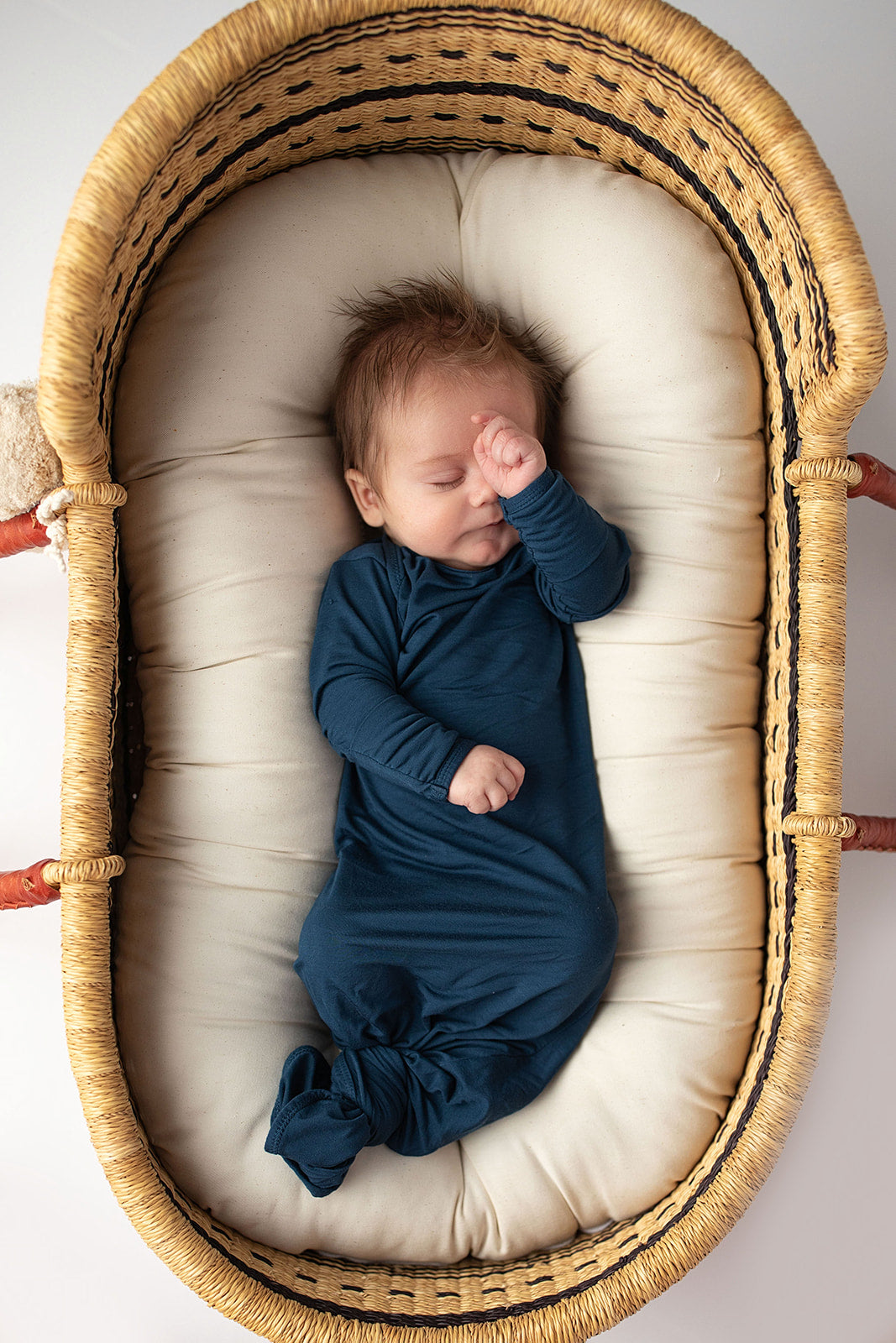 Three Little Tots Navy Blue Knotted Baby Gown. Ultra soft number 1 knotted newborn baby gown. Newborn photoshoot idea or for a pregnancy announcement. Newborn baby in a Snuggle Me Organics in a Plum + Sparrow Moses Basket 