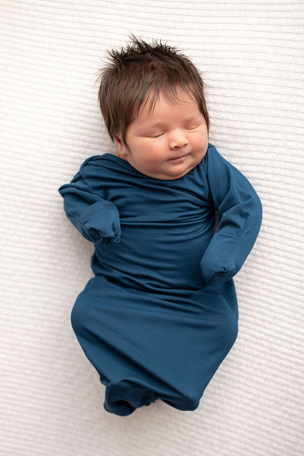 Three Little Tots Navy Blue Knotted Baby Gown. Ultra soft number 1 knotted newborn baby gown. Newborn photoshoot idea or for a pregnancy announcement. Newborn baby in a Snuggle Me Organics in a Plum + Sparrow Moses Basket 