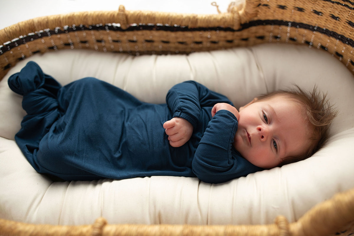 Three Little Tots Navy Blue Knotted Baby Gown. Ultra soft number 1 knotted newborn baby gown. Newborn photoshoot idea or for a pregnancy announcement. Newborn baby in a Snuggle Me Organics in a Plum + Sparrow Moses Basket 