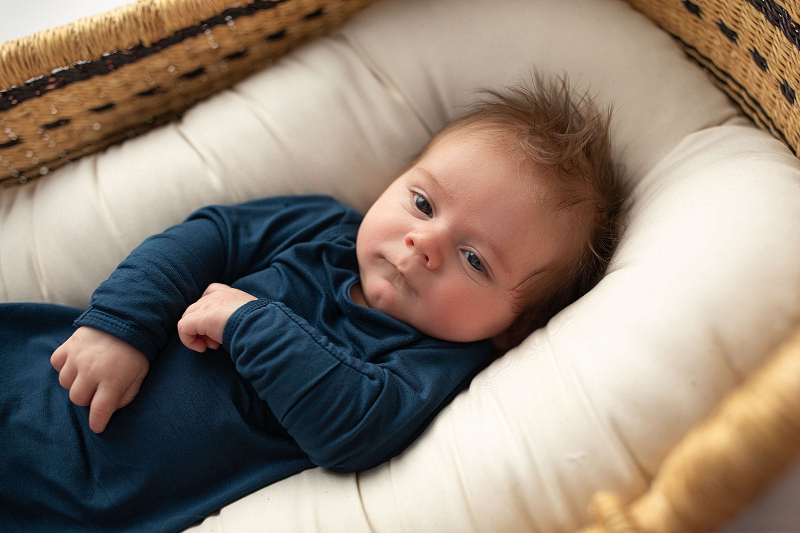 Three Little Tots Navy Blue Knotted Baby Gown. Ultra soft number 1 knotted newborn baby gown. Newborn photoshoot idea or for a pregnancy announcement. Newborn baby in a Snuggle Me Organics in a Plum + Sparrow Moses Basket 