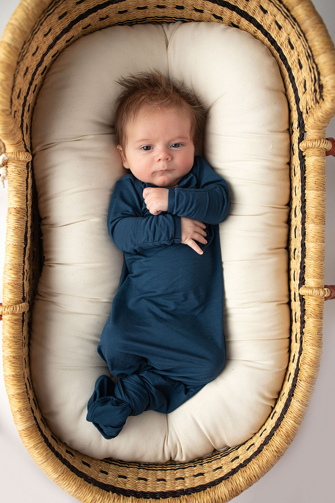 Three Little Tots Navy Blue Knotted Baby Gown. Ultra soft number 1 knotted newborn baby gown. Newborn photoshoot idea or for a pregnancy announcement. Newborn baby in a Snuggle Me Organics in a Plum + Sparrow Moses Basket 