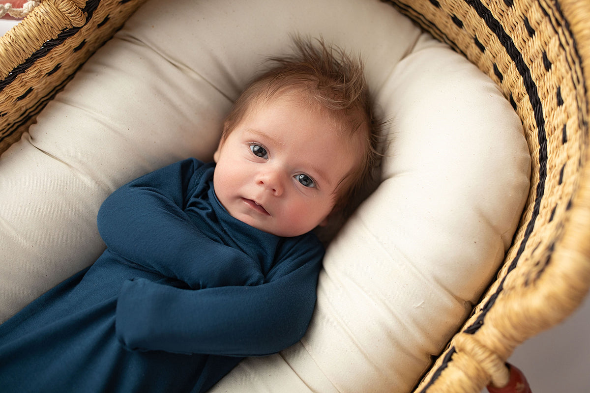 Three Little Tots Navy Blue Knotted Baby Gown. Ultra soft number 1 knotted newborn baby gown. Newborn photoshoot idea or for a pregnancy announcement. Newborn baby in a Snuggle Me Organics in a Plum + Sparrow Moses Basket 
