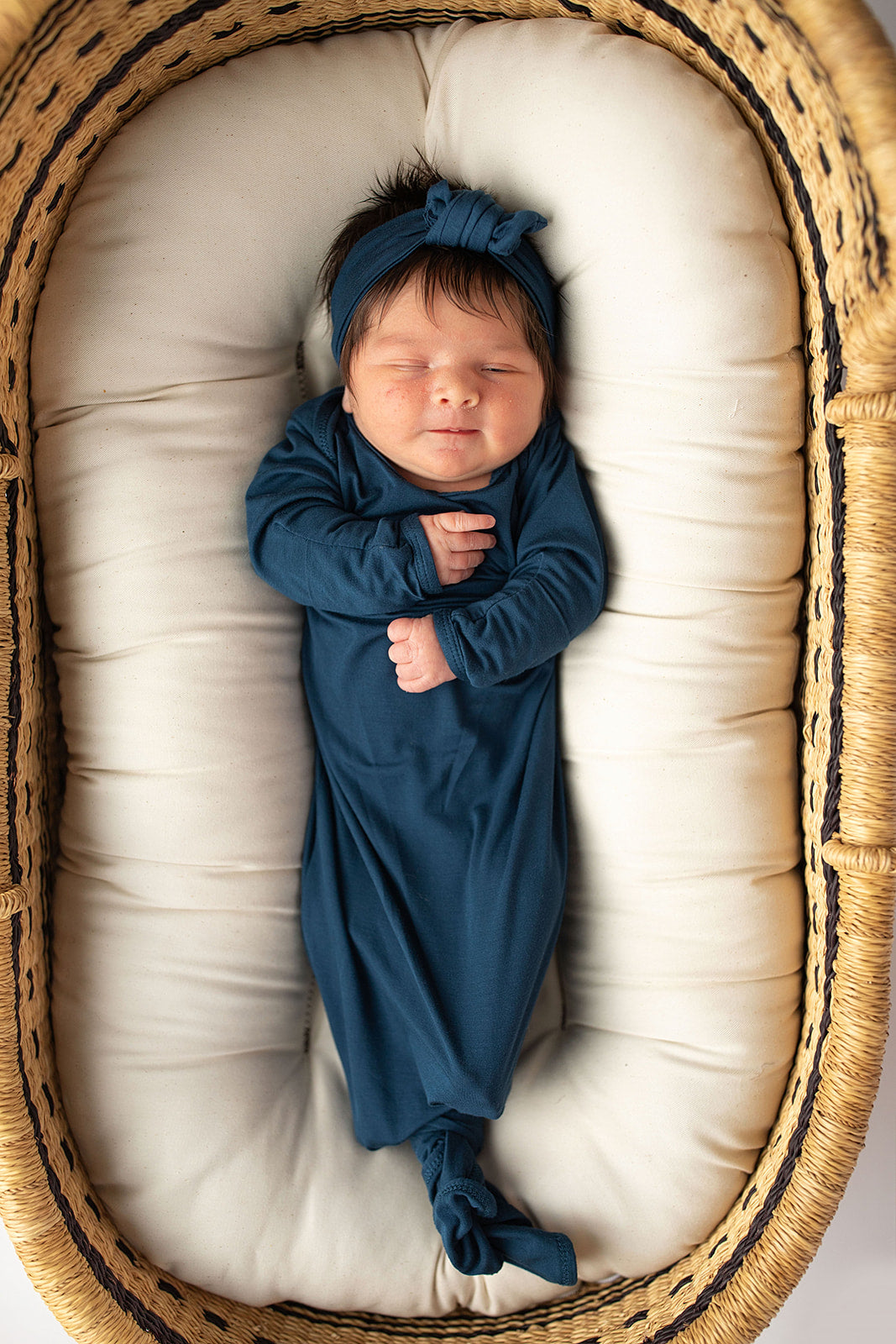 Three Little Tots Navy Blue Knotted Baby Gown. Ultra soft number 1 knotted newborn baby gown. Newborn photoshoot idea or for a pregnancy announcement. Newborn baby in a Snuggle Me Organics in a Plum + Sparrow Moses Basket 