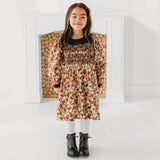 Young girl wearing a floral dress standing in a minimalistic room with wooden logs in the background.