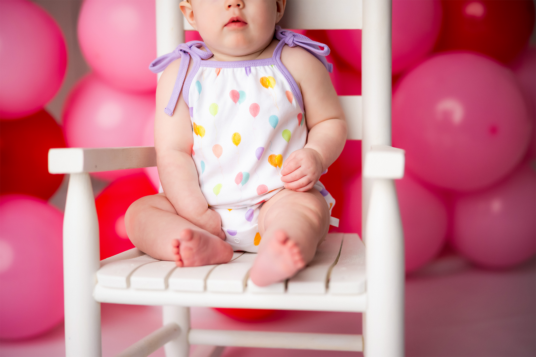 Baby sitting on a white chair surrounded by pink balloons