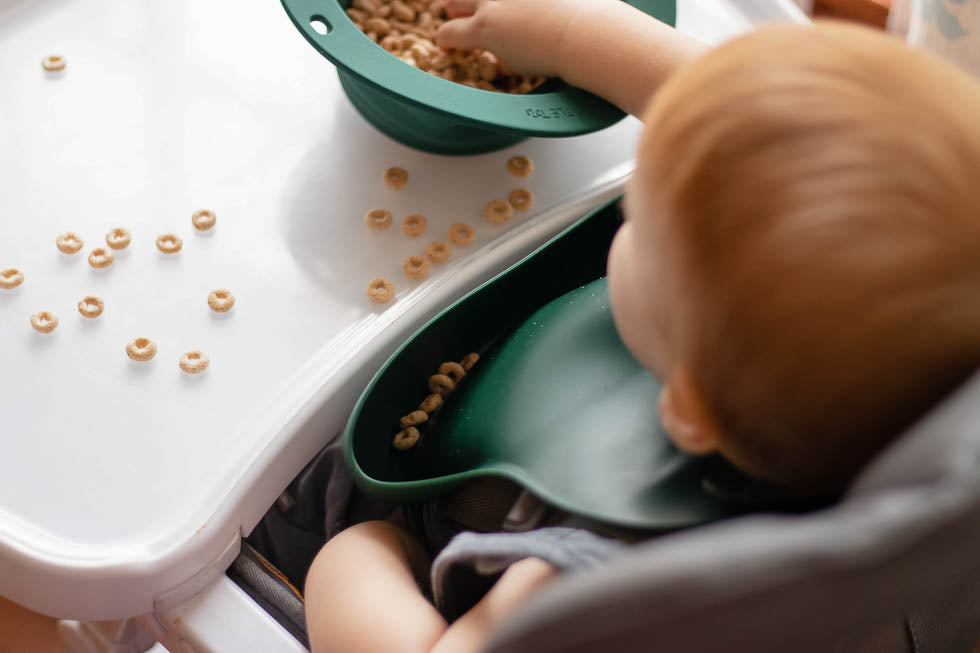 Little boy eating Cheerios. Designed in the USA, Three Little Tots food-grade silicone bibs with crumb catcher ensure comfort and cleanliness for your baby.