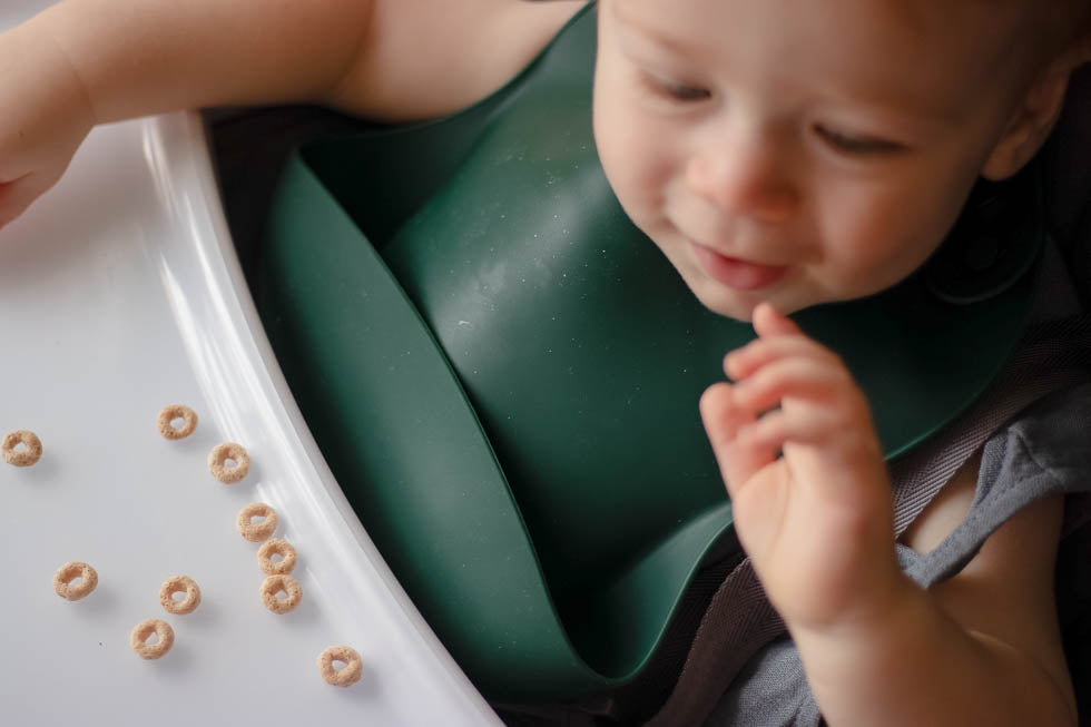 Eating Cheerios in a high chair. Designed in the USA, Three Little Tots food-grade silicone bibs with crumb catcher ensure comfort and cleanliness for your baby.