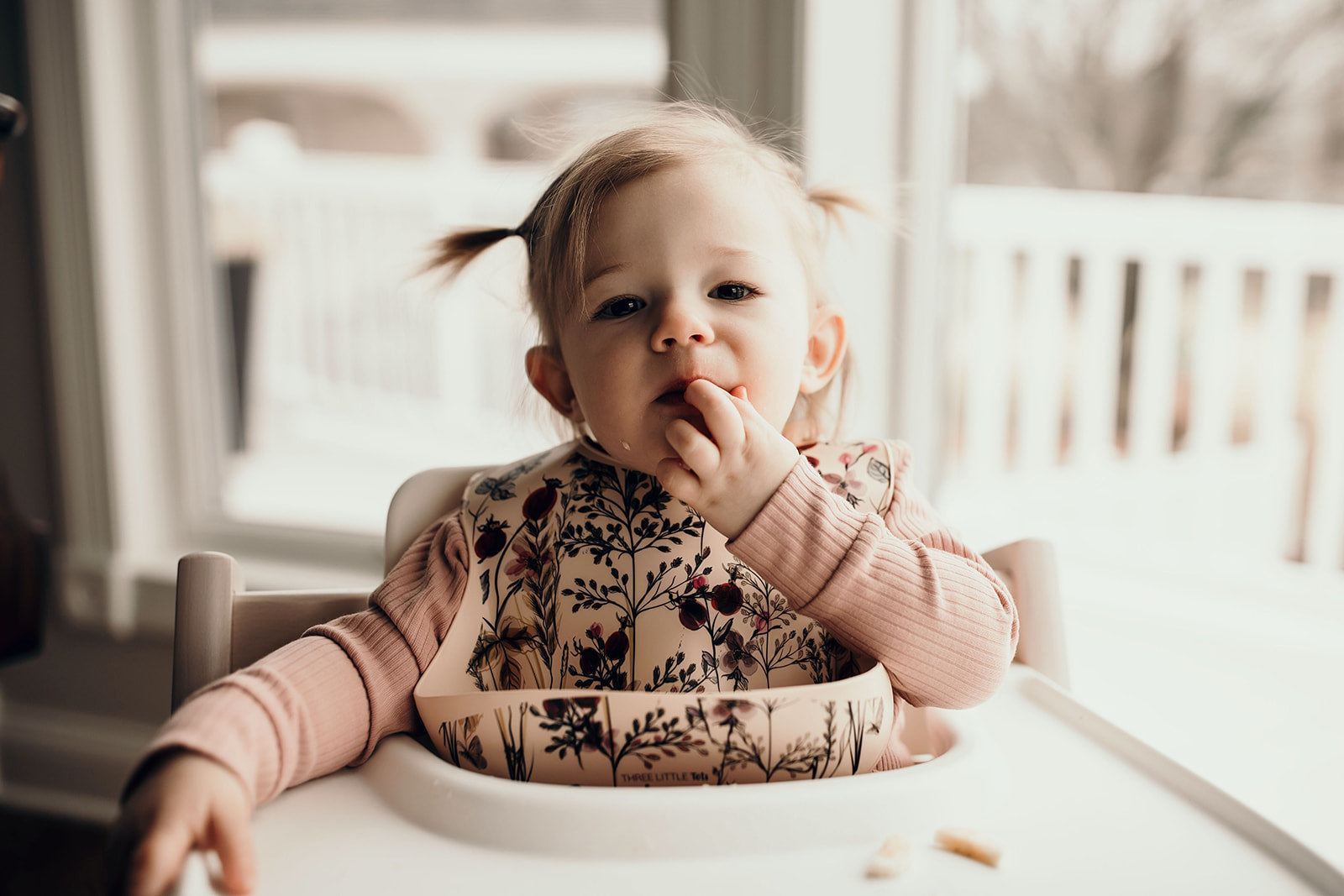 Little girl eating puffs with a Three Little Tots floral silicone bib