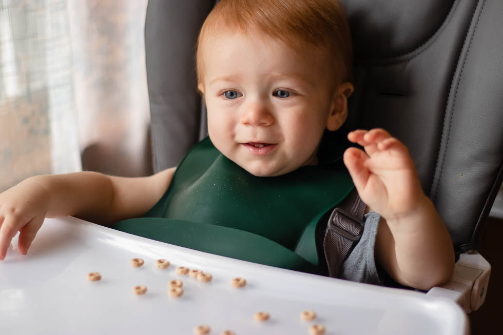 Little Boy in highchair eating Cheerios while wearing a Three Little Tots Bib. Designed in the USA, Three Little Tots food-grade silicone bibs with crumb catcher ensure comfort and cleanliness for your baby