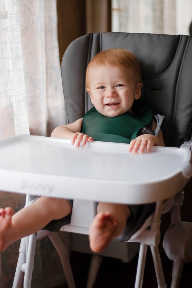 Toddler boy in high chair. Designed in the USA, Three Little Tots food-grade silicone bibs with crumb catcher ensure comfort and cleanliness for your baby.