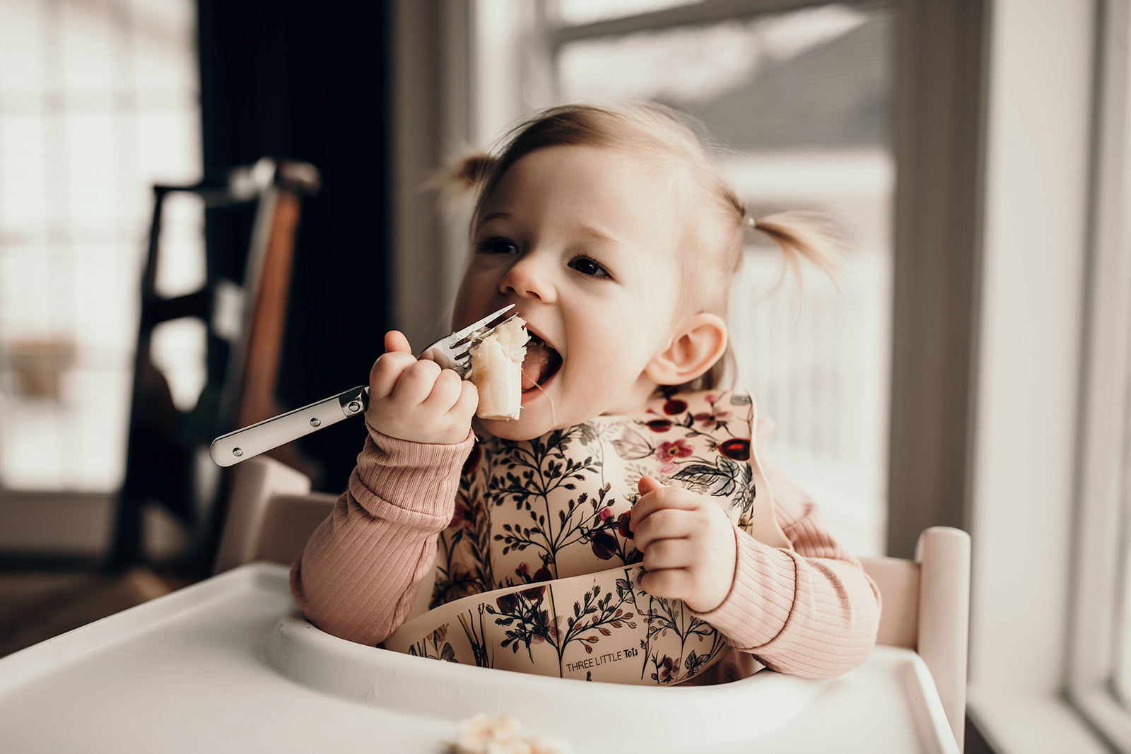 Little girl eating smashed bananas with a Three Little Tots floral silicone bib