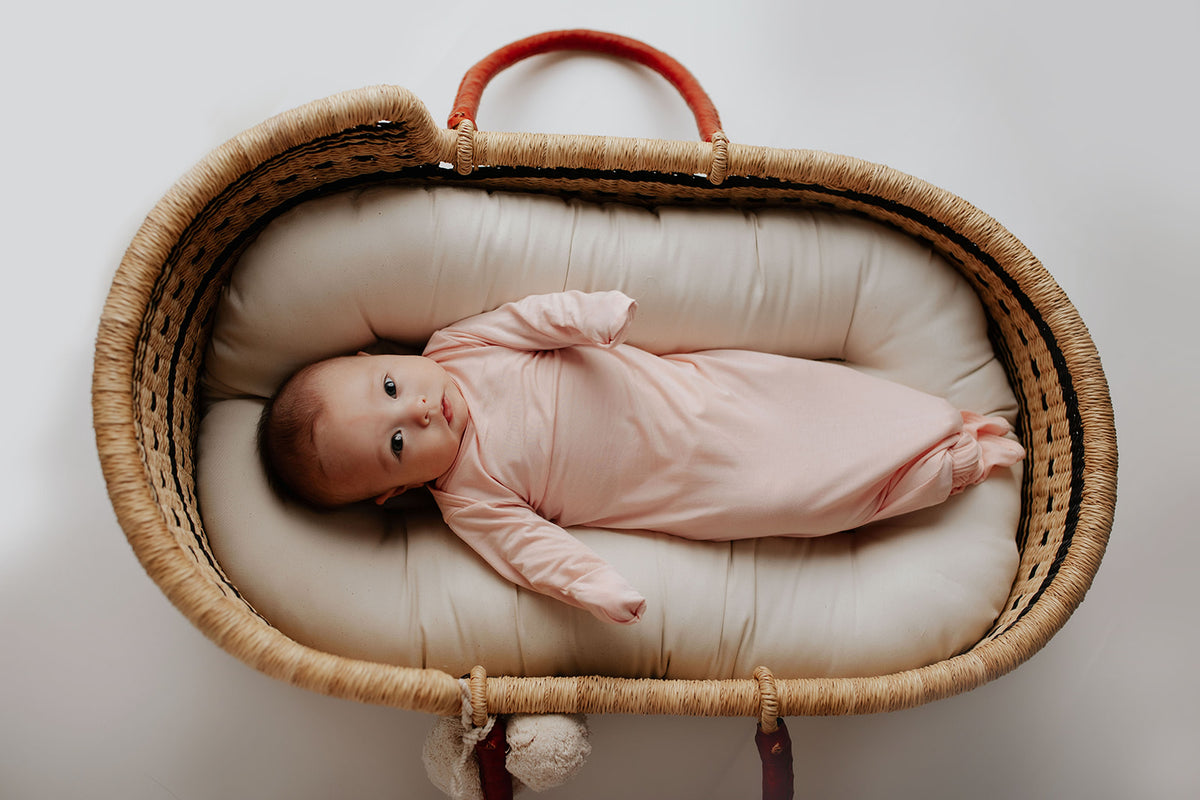 Newborn baby in a Three Little Tots Knotted Baby gown. Baby is laying in a Snuggle Me Organics inside a Plum and Sparrow Moses Basket
